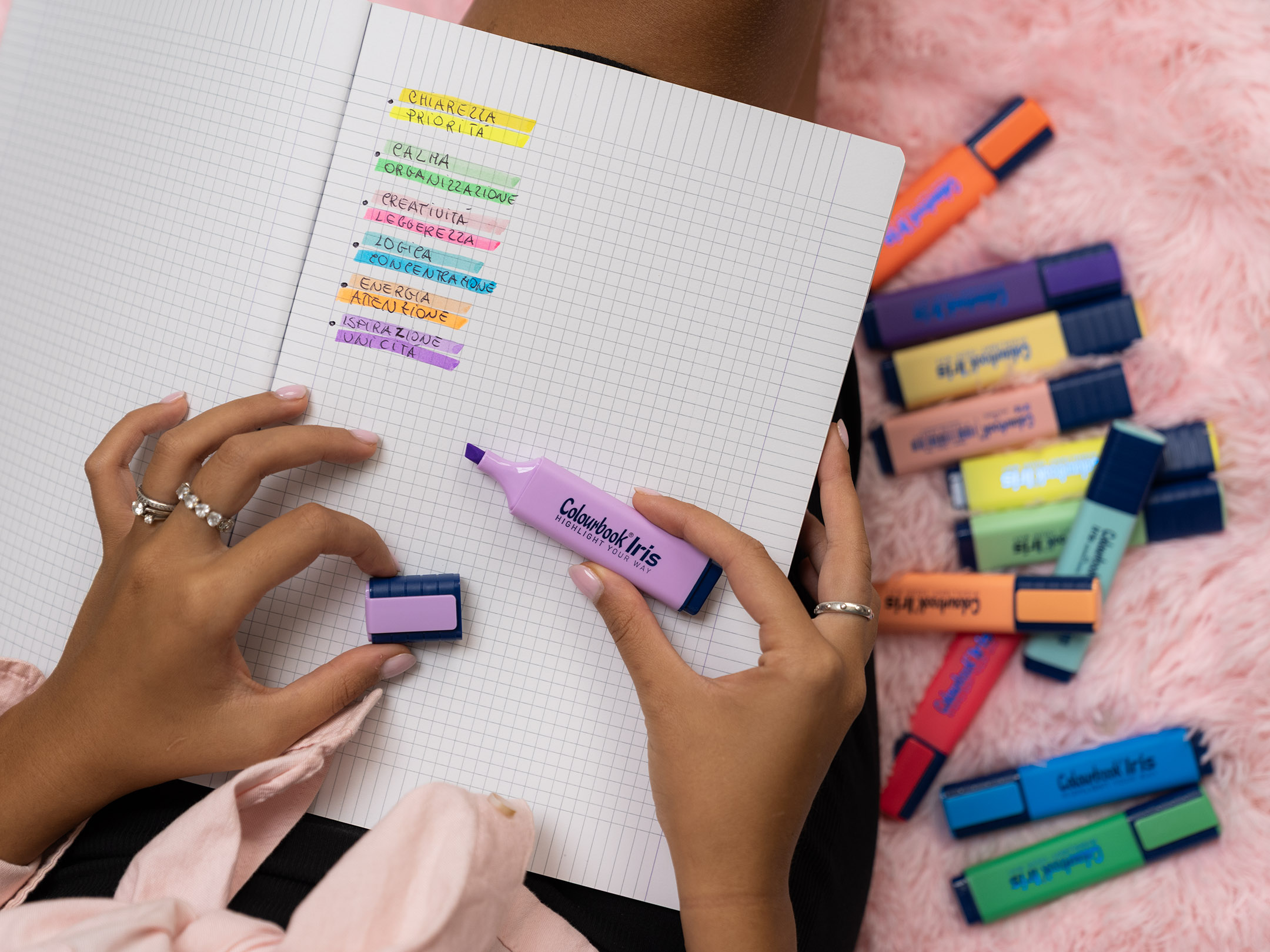 In questa immagine vediamo una mano che utilizza un evidenziatore lilla Colourbook Iris su un quaderno a quadretti. Le righe sono organizzate con diversi colori, ognuno associato a una parola chiave come chiarezza, priorità, creatività, energia o ispirazione. Attorno al quaderno sono sparsi altri evidenziatori Colourbook in vivaci tonalità pastello e fluo. L’insieme mette in risalto l’uso del color code: ogni colore diventa un segnale visivo che aiuta a distinguere concetti, organizzare idee e dare più energia allo studio e al lavoro.