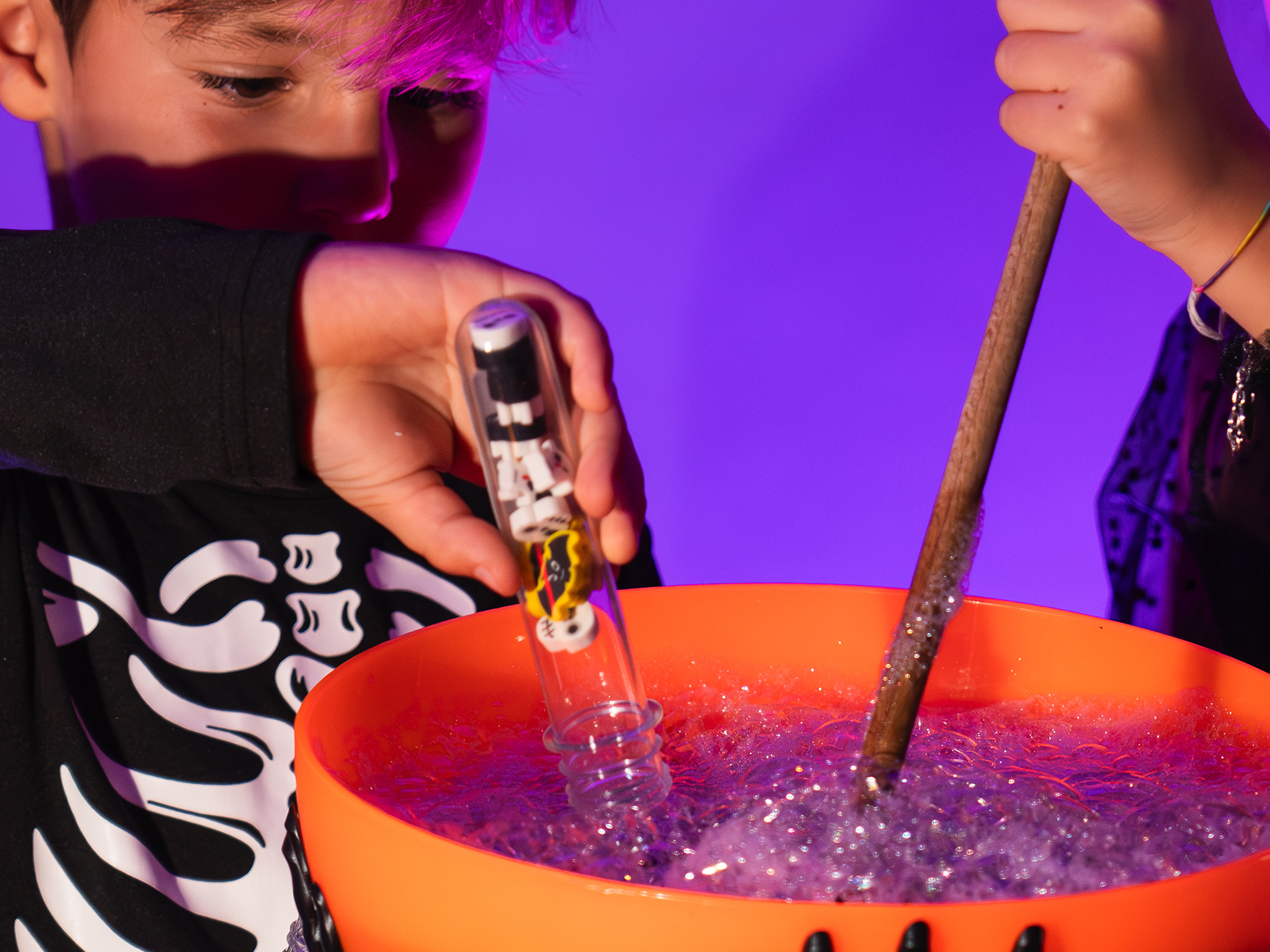 Un bambino vestito da scheletro gioca con una zucca arancione piena di bolle scintillanti, mescolandole con un bastoncino come in un incantesimo. Nella mano tiene una provetta Colourbook con piccole decorazioni a tema Halloween. La scena, illuminata da luce viola e calda, trasmette un senso di magia e divertimento tipico della notte delle streghe.
